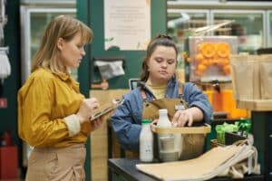 Young woman with Down syndrome training in supermarket and helping customers with social worker assisting, ABA Therapy