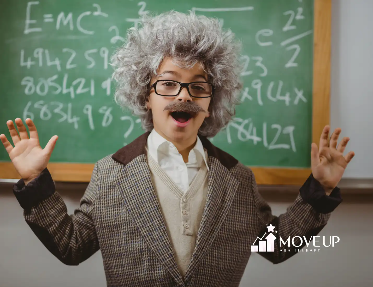 A little boy cosplaying Einstein smiling in front of chalkboard