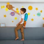 Young boy wearing a toy jetpack, standing on a couch and reaching for a cardboard rocket.