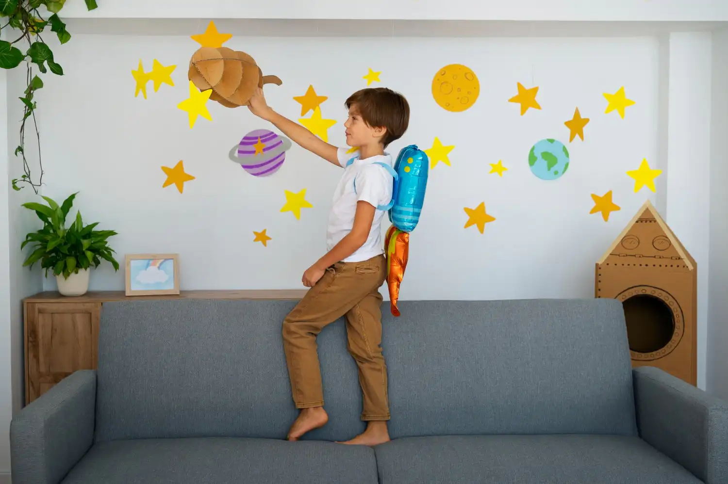 Young boy wearing a toy jetpack, standing on a couch and reaching for a cardboard rocket.