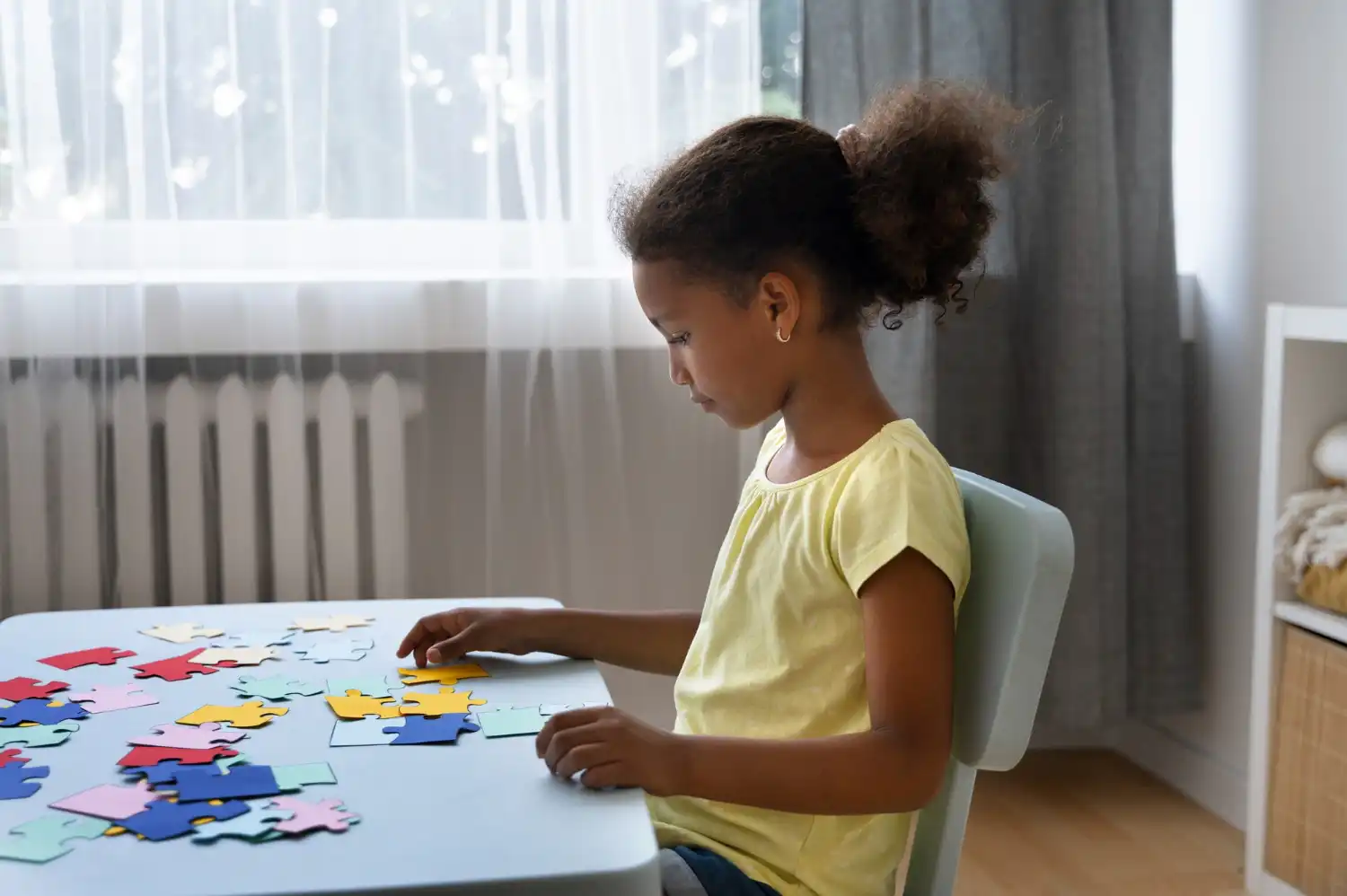 A young girl in a yellow shirt sits at a table, focused on assembling a colorful puzzle.