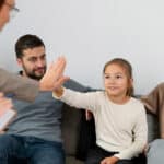 Young girl giving a high-five to ABA therapist while sitting on a couch with her smiling parents.