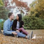 Autistic couple sitting on autumn leaves, smiling at each other under a large tree at a park in Maryland.