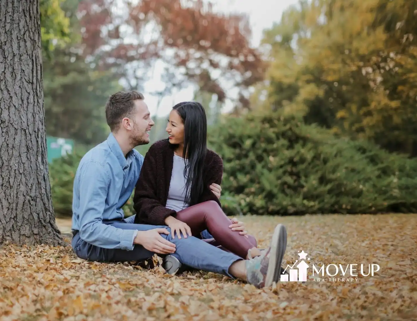 Autistic couple sitting on autumn leaves, smiling at each other under a large tree at a park in Maryland.