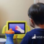 A child with short dark hair sits at a table watching a tablet with a yellow case. In front is a sippy cup and a bowl of fruit.