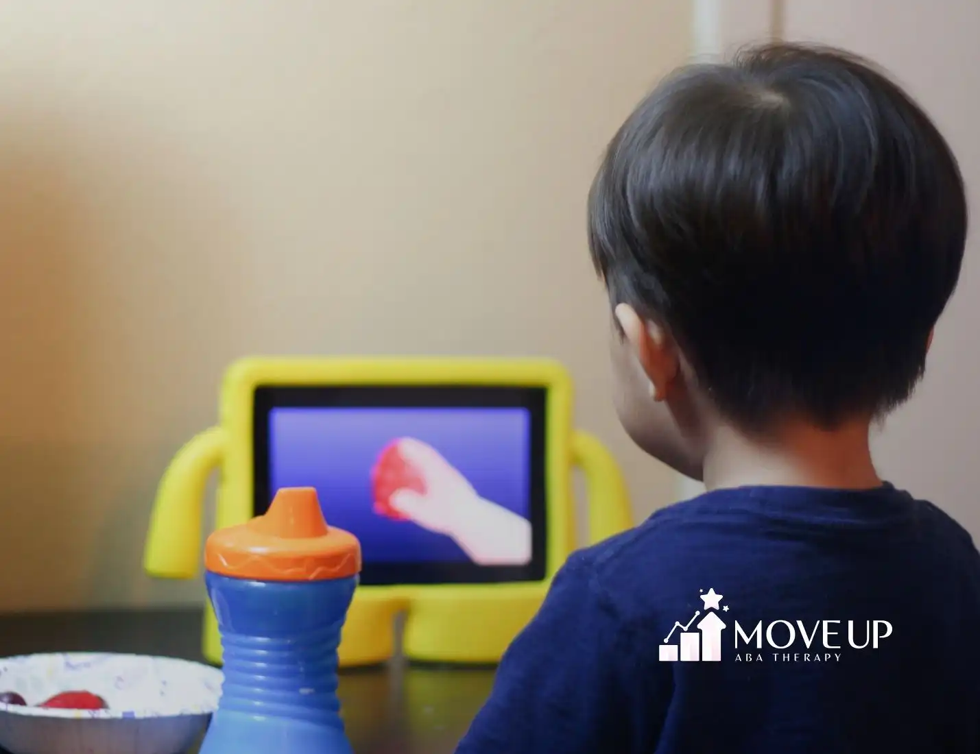 A child with short dark hair sits at a table watching a tablet with a yellow case. In front is a sippy cup and a bowl of fruit.