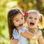 Two young girls hug outdoors in warm sunlight, laughing joyfully; one wears a blue dress, the other a yellow top with a red flower in her hair.