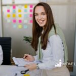 A BCBA smiling with BCBA task list clipboard in an office while colleague uses phone nearby in MD.