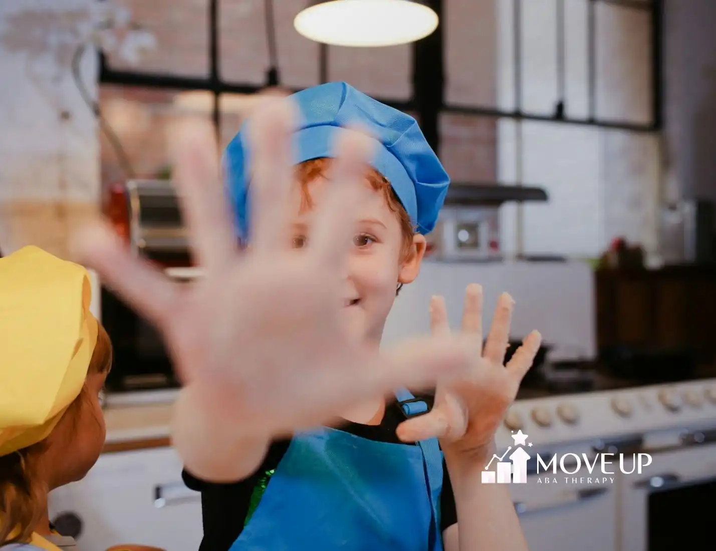 An autistic child in a chef hat holding up flour-covered hands in a kitchen during ABA therapy session in MD.