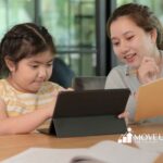 A young girl with braids focuses on a tablet, while a smiling woman holds a notebook and pencil beside her.