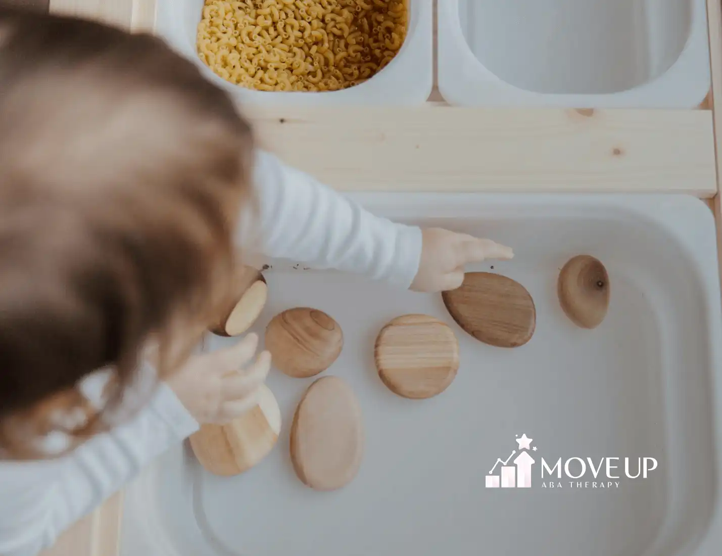 An autistic toddler pointing at smooth wooden sensory stones in a white sensory bin during ABA therapy session in Maryland.