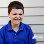 A young boy in a blue shirt makes a playful, scrunched face against a beige siding background.