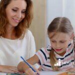 Female ABA therapist woman helping autistic girl with braids draw using colored pencils in ABA therapy.