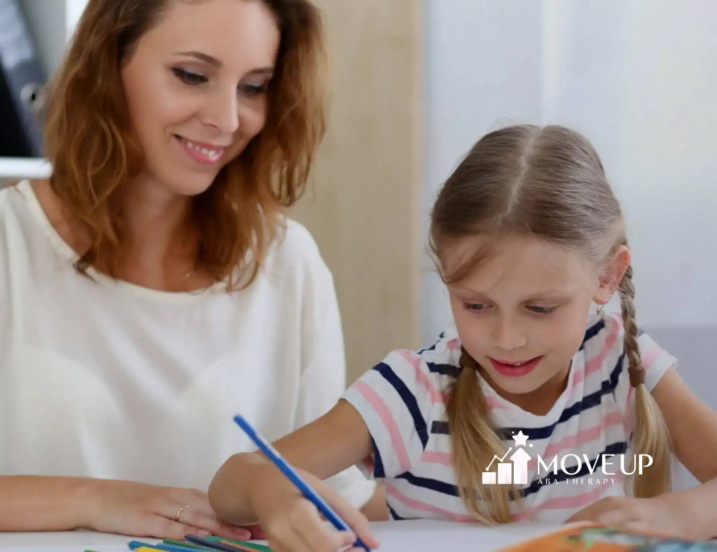 Female ABA therapist woman helping autistic girl with braids draw using colored pencils in ABA therapy.