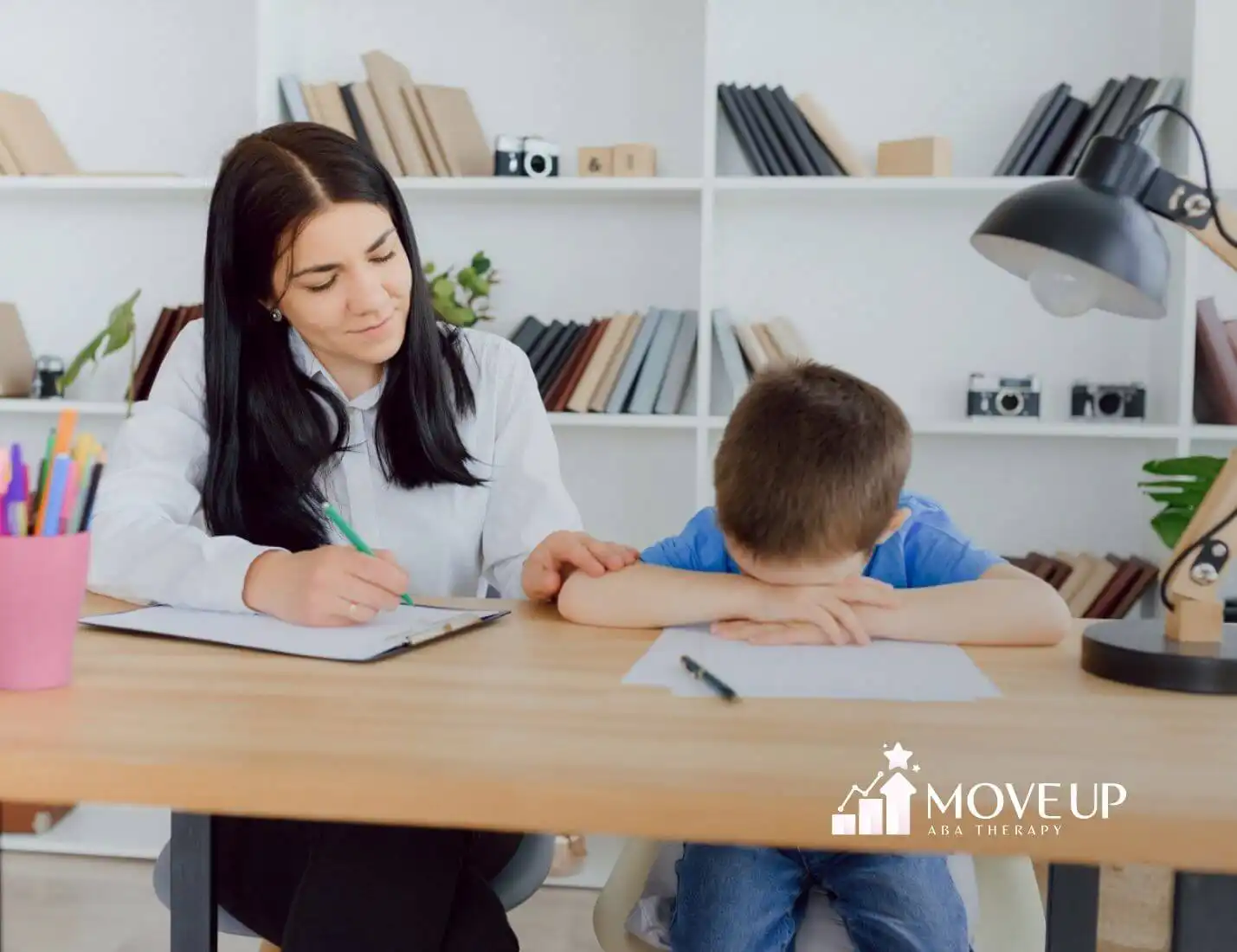 ABA therapist comforting autistic with head down at desk during ABA therapy session.