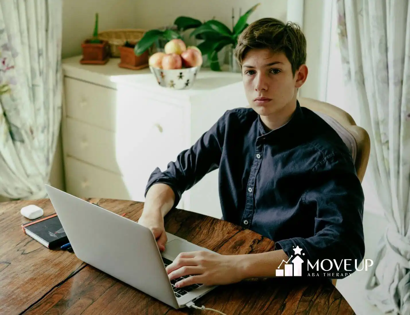 An autistic teen in blue shirt using laptop at home table with phone, notebook, and earbuds nearby after therapy.