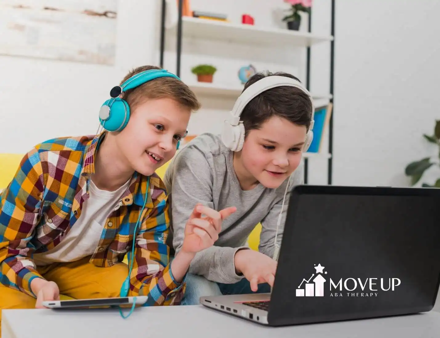 Two autistic boys wearing headphones and smiling while playing on a laptop and tablet in a bright room.