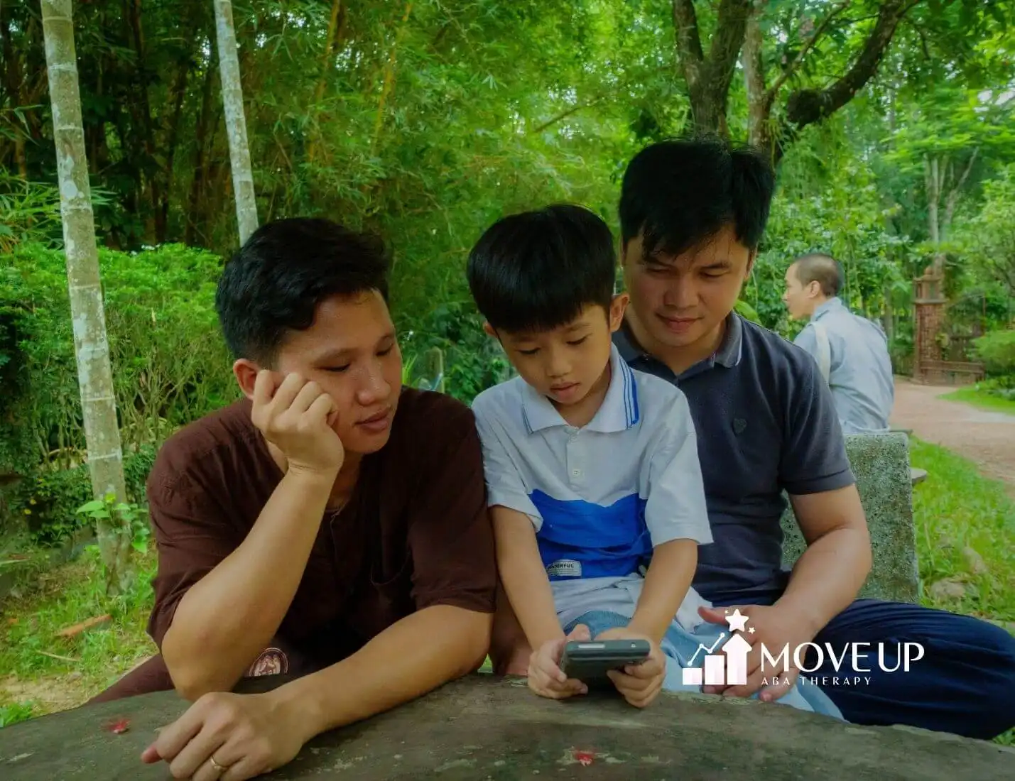Autistic child shows phone after therapy to two adults on a bench in a green park; all focused on screen.