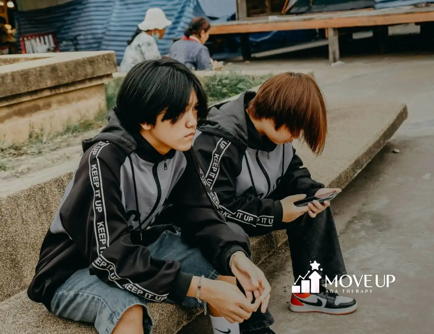 Two autistic teens in matching hoodies sitting on outdoor steps, one using a smartphone after ABA therapy.