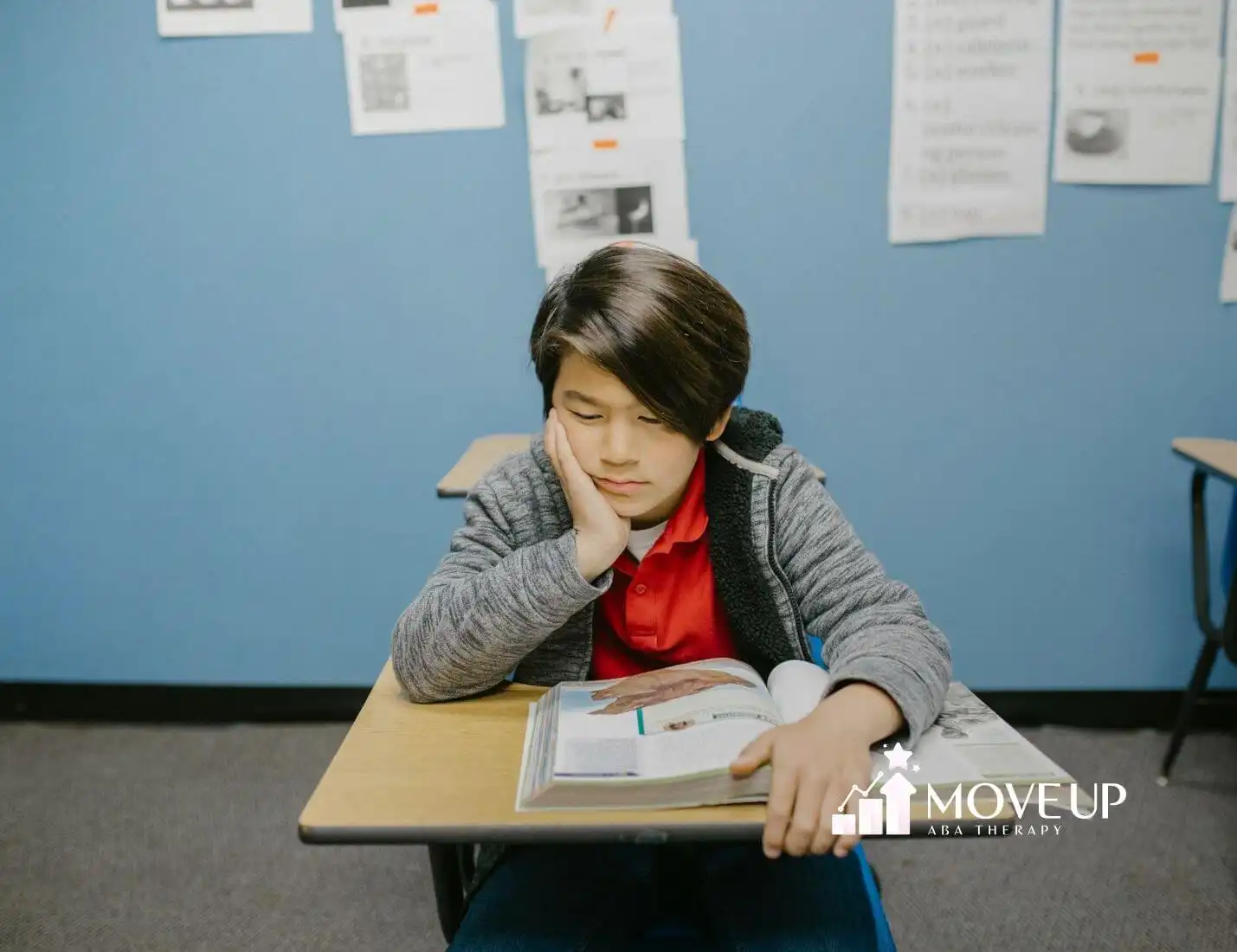 Autistic young boy in red shirt reading a book at his desk in a blue-walled classroom after ABA therapy.
