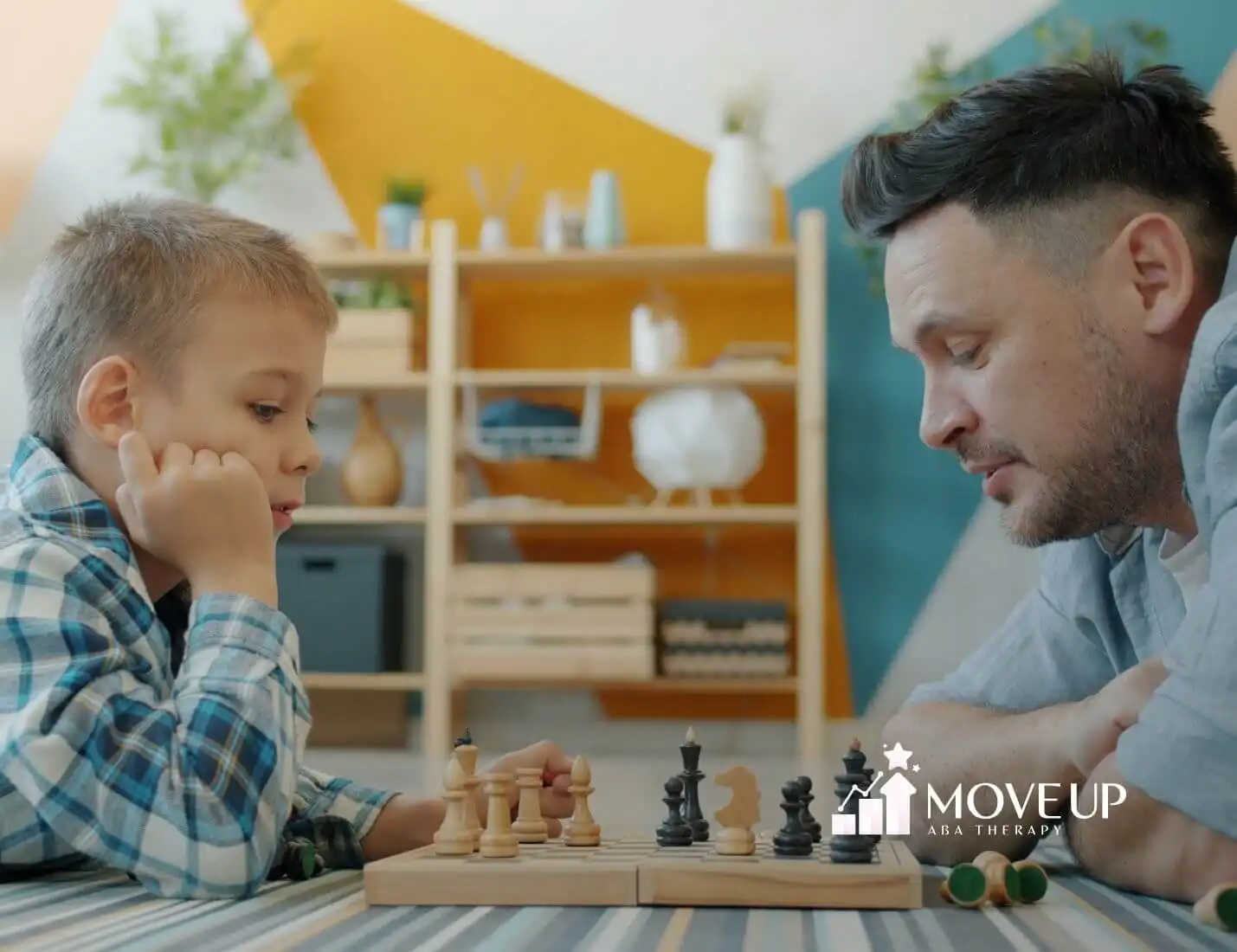 Male therapist and autistic boy lying on the floor playing chess together in a cozy living room after ABA therapy.