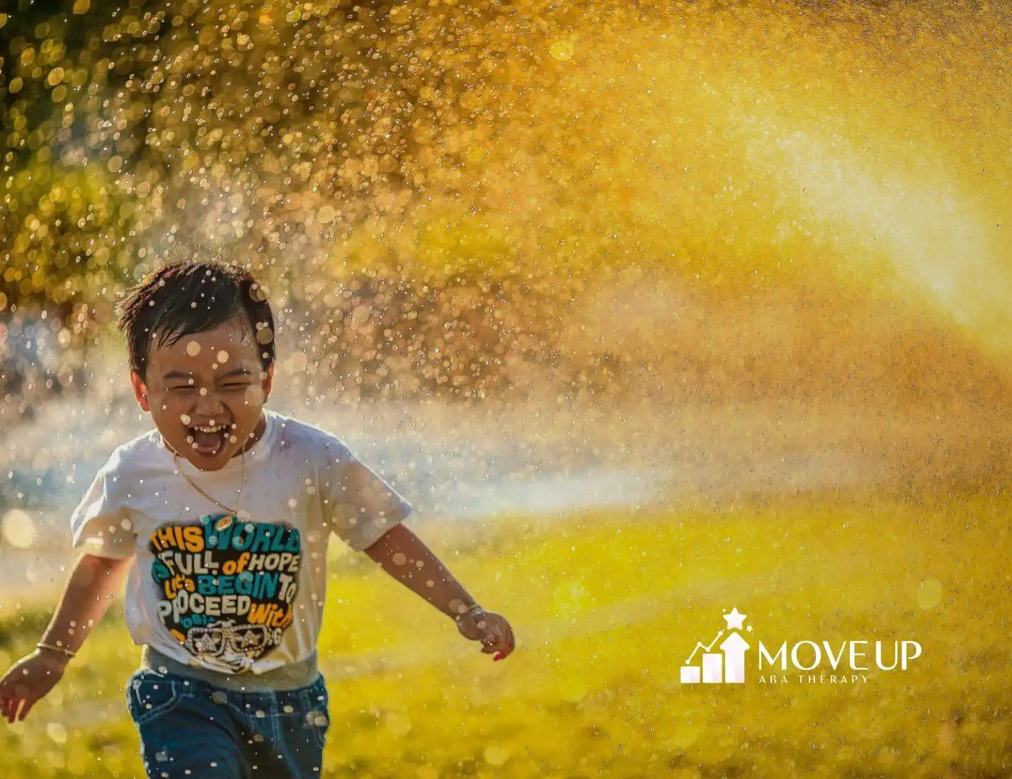 Autistic young boy runs through sprinkler spray in sunlight, wearing white shirt and blue shorts after ABA therapy.