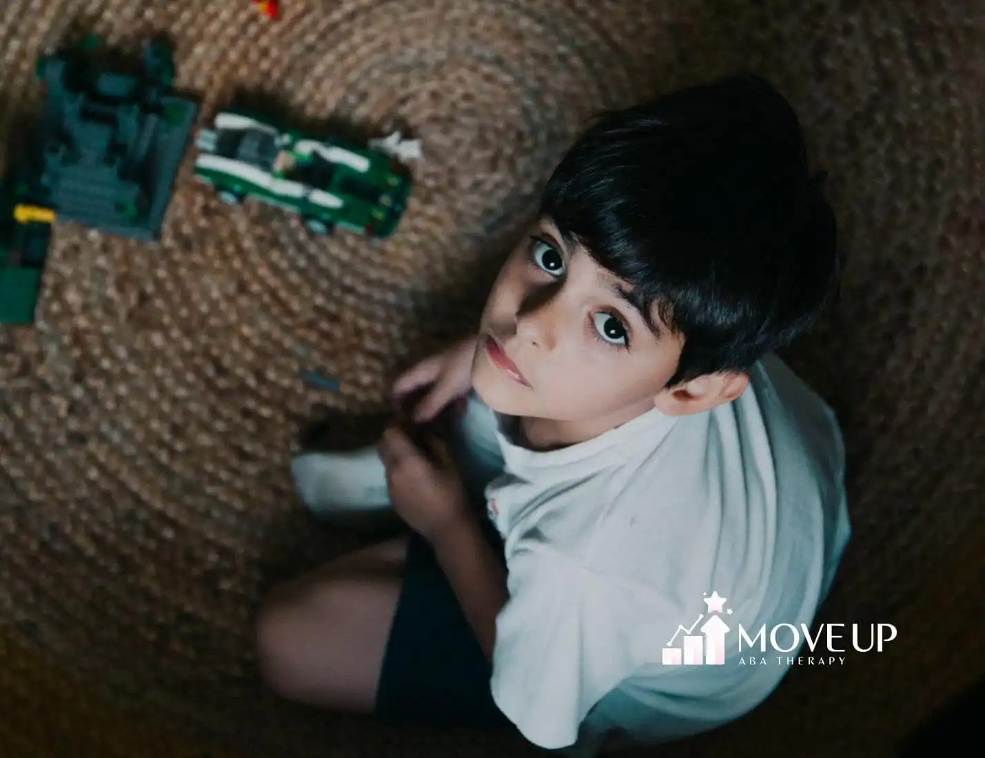 A young boy with autism sitting on a woven rug, surrounded by LEGO toys, looking up at the camera with big, curious eyes.