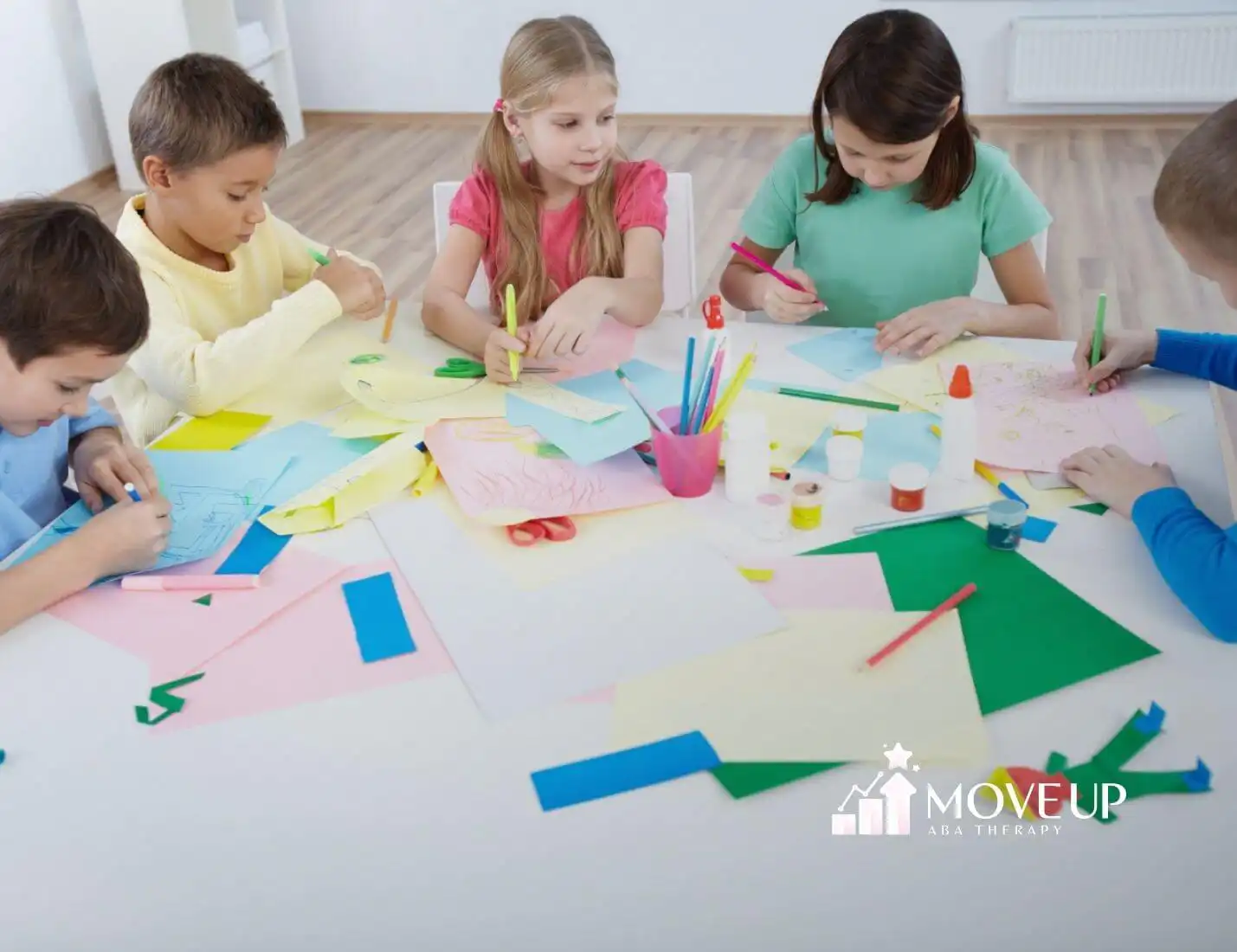 Five children with autism sitting at a table working on a colorful paper craft project.