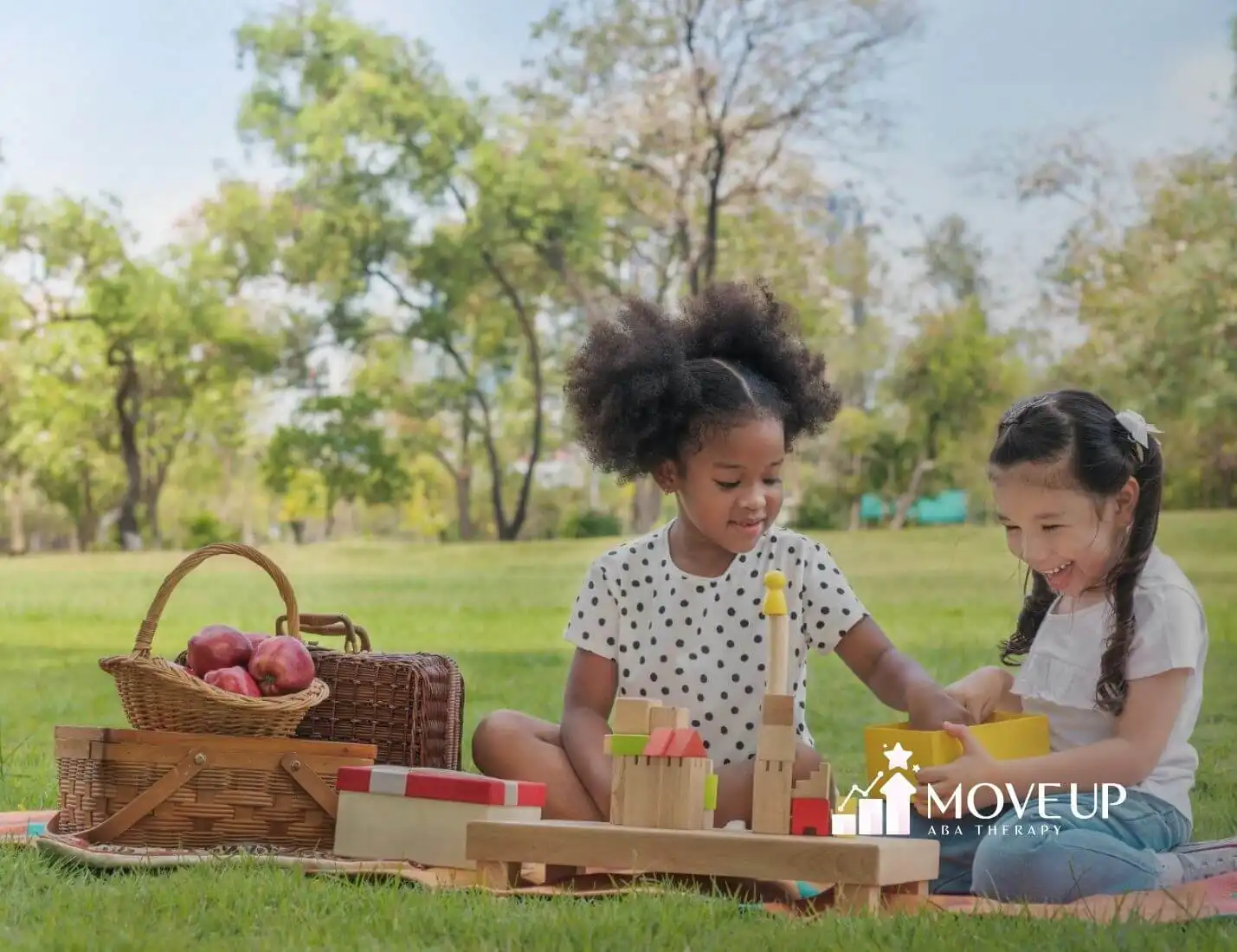 Two young girls with autism playing with wooden blocks on a picnic blanket in a sunny outdoor setting.