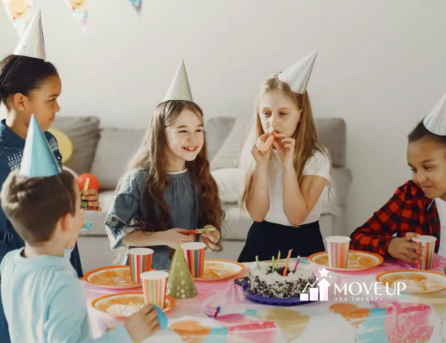 Group of children with autism wearing party hats celebrating a birthday around a table with cake.