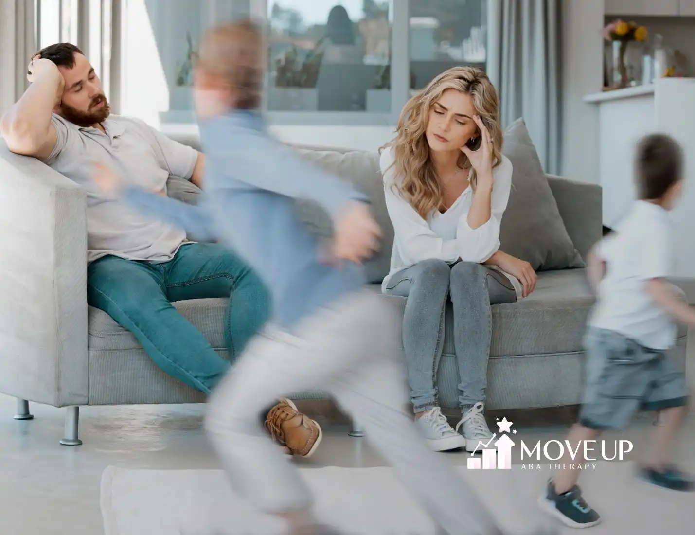 A tired couple sits on a couch, looking stressed, while two blurred children run around them energetically. The scene conveys chaos and exhaustion.