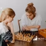 A woman and a young girl focus intently on a chess game at a wooden table. An open wooden box with game pieces is beside them. The mood is calm and thoughtful.