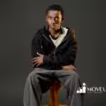 A young person in casual attire, wearing a black hoodie and gray jeans, sits on a wooden stool against a dark background. Their expression is calm and contemplative.