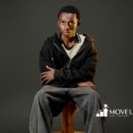 A young person in casual attire, wearing a black hoodie and gray jeans, sits on a wooden stool against a dark background. Their expression is calm and contemplative.