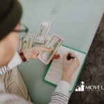 A person is sitting on a pastel blue couch, holding U.S. dollar bills in one hand and writing on a planner with the other. The atmosphere is organized and focused.