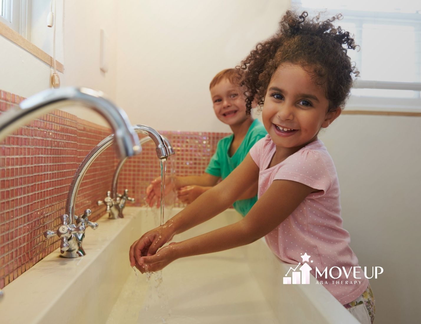 Two children smiling while washing their hands in a brightly lit bathroom. The girl wears pink and the boy green. The atmosphere is cheerful and playful.