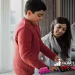 A boy in a red sweater plays with toy trains on a table, while a woman, smiling, watches from a sofa. The scene is indoors with a relaxed and warm atmosphere.