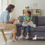 Therapist with a clipboard sitting and talking with a young girl who is holding a large teddy bear on a couch in a bright living room