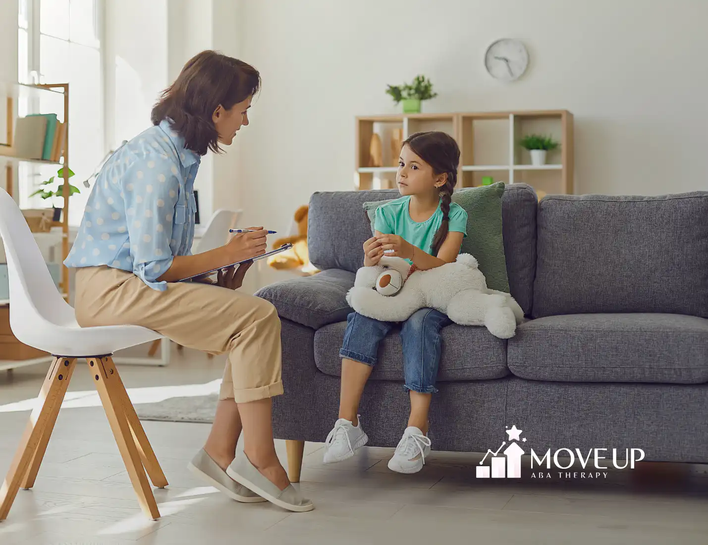 Therapist with a clipboard sitting and talking with a young girl who is holding a large teddy bear on a couch in a bright living room
