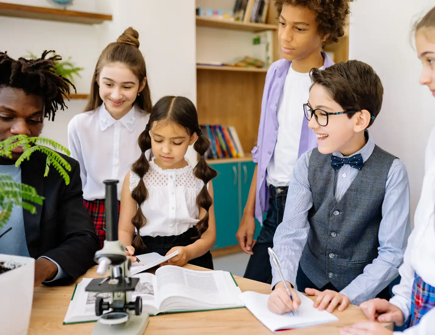 A teacher and a group of school children gathered around a table with a microscope, books, and notebooks during a science lesson in a classroom.