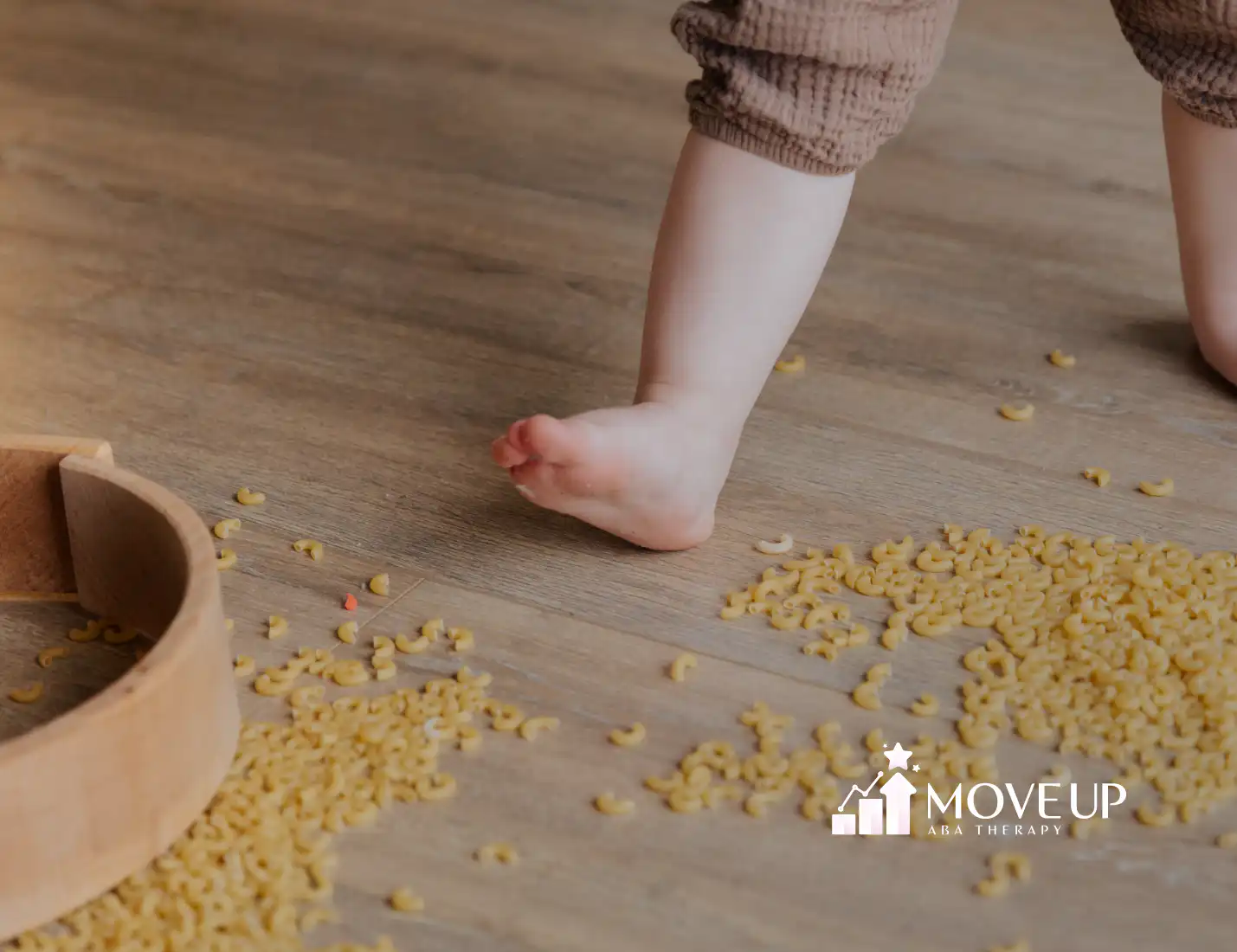 A barefoot toddler standing on a wooden floor scattered with dry macaroni pasta, with a wooden bowl nearby.