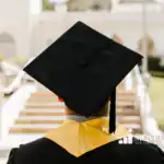 Rear view of a graduate in a black cap and gown with a gold stole, walking up outdoor stone steps toward a sunlit building.