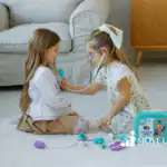 Two young girls sitting on a rug playing doctor, one holding a toy stethoscope to the other's chest, with a play medical kit spread out around them.