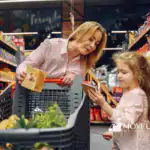 Mother and young daughter grocery shopping together, with the child holding a notepad shopping list while her mom shows her a food item in the store aisle