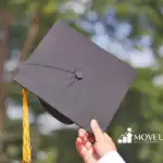 Hand holding up a black graduation cap with a gold tassel against a blurred outdoor background of trees.
