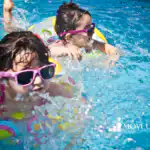 Two children in pink sunglasses splashing in a pool on colorful inflatable rings