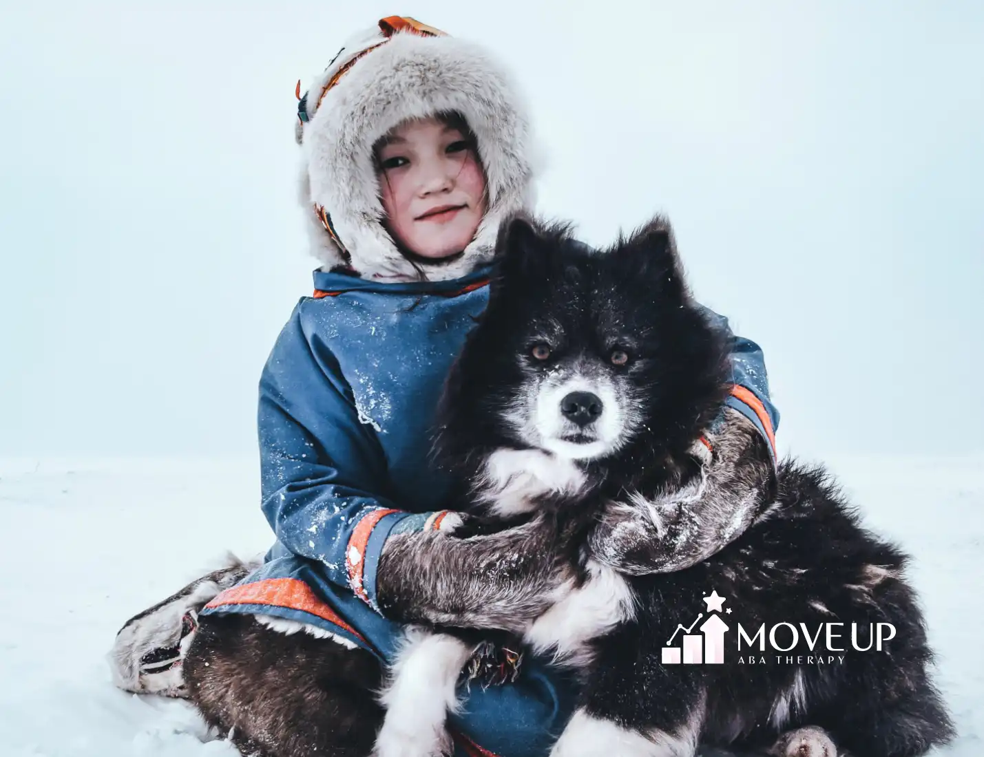 Child in a fur-lined winter hood and blue coat sitting in the snow, hugging a black and white dog.