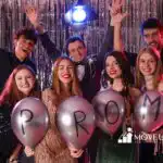 A group of four friends in formal attire smiling at a prom party, with colorful decorations in the background.