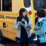 Two students in uniforms holding books and talking beside a yellow school bus